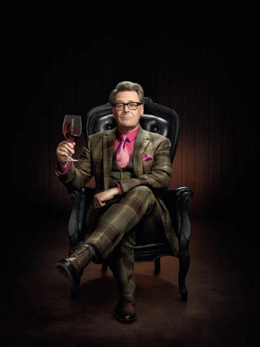 Middle-aged man in a plaid suit holding a wine glass seated in a black leather armchair against a dark background