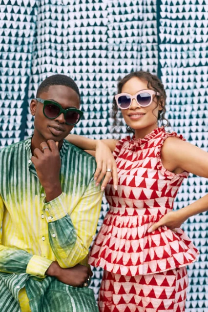 Two people in sunglasses and colorful outfits pose in front of a triangle-patterned backdrop, showcasing fashion and style.