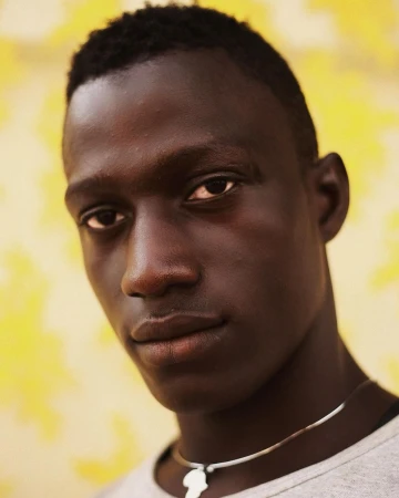 Close-up portrait of a Black man with short dark hair, wearing a silver necklace with an Africa pendant, against a yellow floral background.