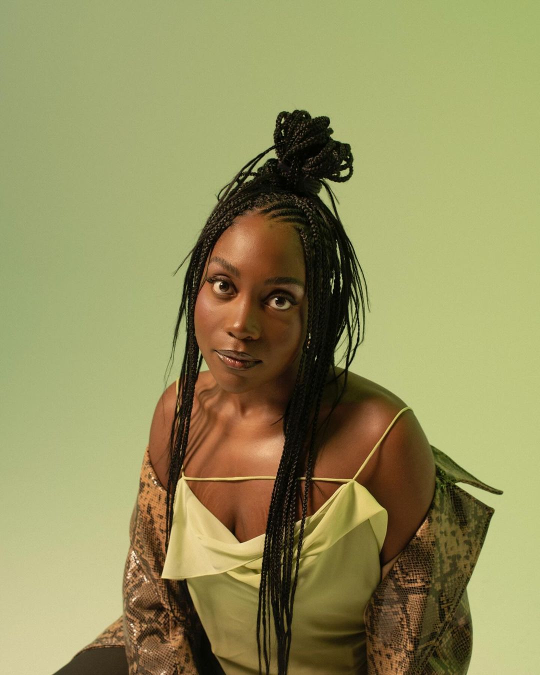 Portrait of a Black woman with box braids wearing a green top and snakeskin jacket against a soft gradient green background.