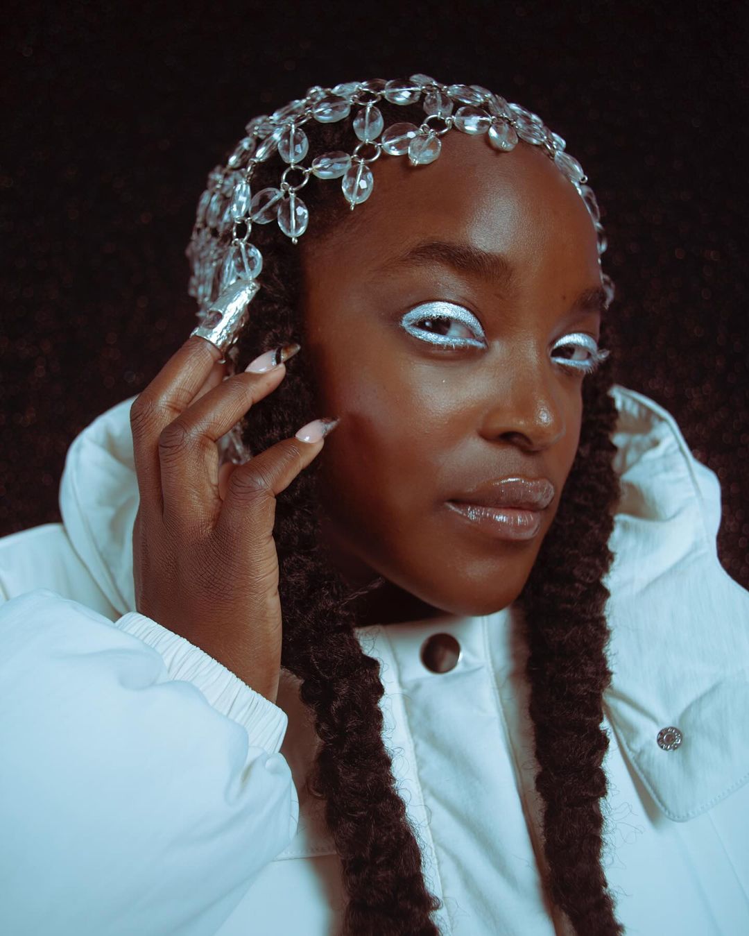 Close-up of a Black woman with an embellished headpiece and white eye makeup wearing a puffy white coat against a dark backdrop.