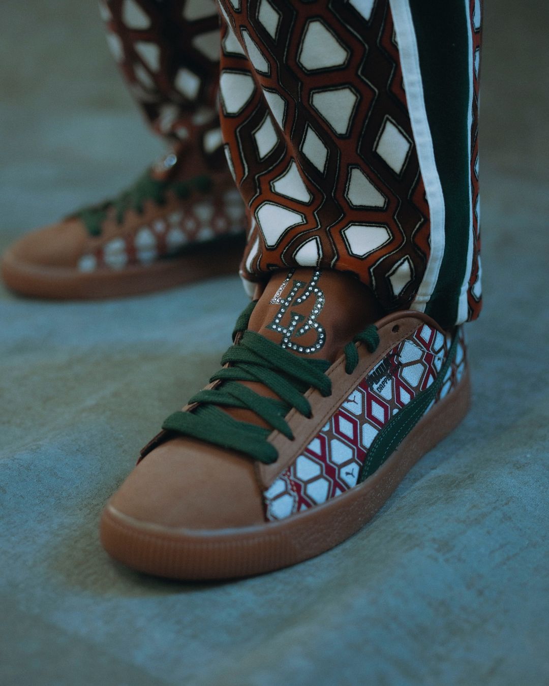 Close-up of brown Puma sneakers with green laces, showcasing a geometric pattern design on the side and tongue, paired with patterned pants.