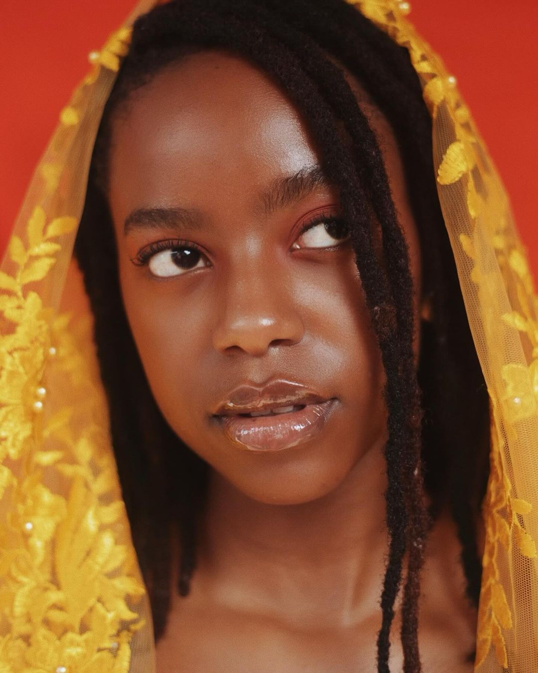 Portrait of a dark-skinned woman with dreadlocks, wearing a yellow floral lace veil, against a solid orange background, looking up.