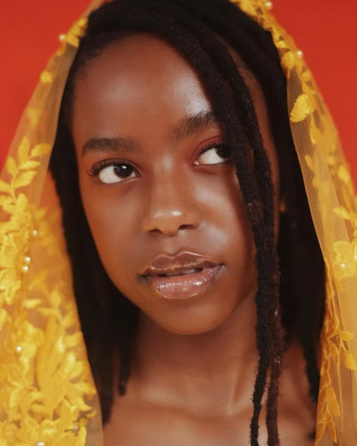 Portrait of a dark-skinned woman with dreadlocks, wearing a yellow floral lace veil, against a solid orange background, looking up.