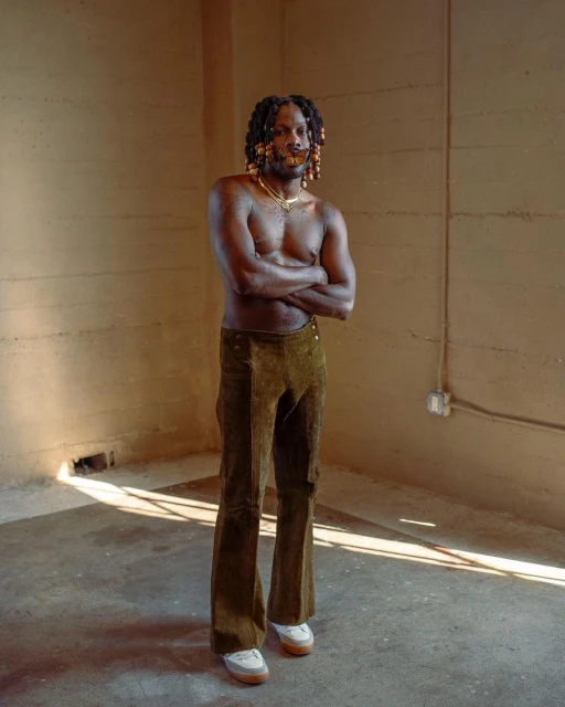 Full-length portrait of a shirtless Black man in bell-bottoms and sneakers standing indoors against a tan wall with his arms crossed.