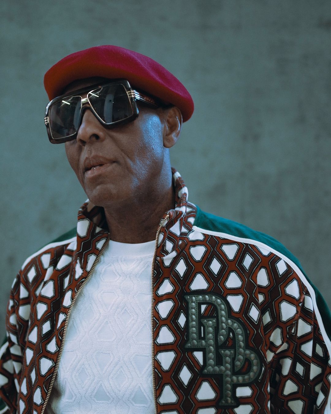 Stylish man with a red beret, sunglasses, and patterned jacket, posing in an indoor setting with a concrete-like background, close-up shot.