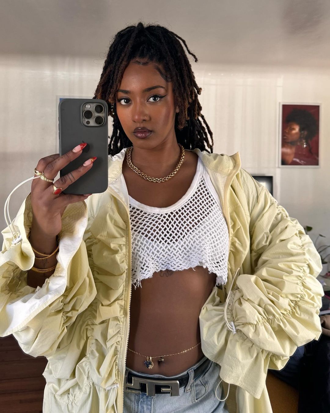 Stylish biracial woman with dreadlocks poses for a selfie in a fashionable outfit with a white crop top, a jacket, and jewelry.