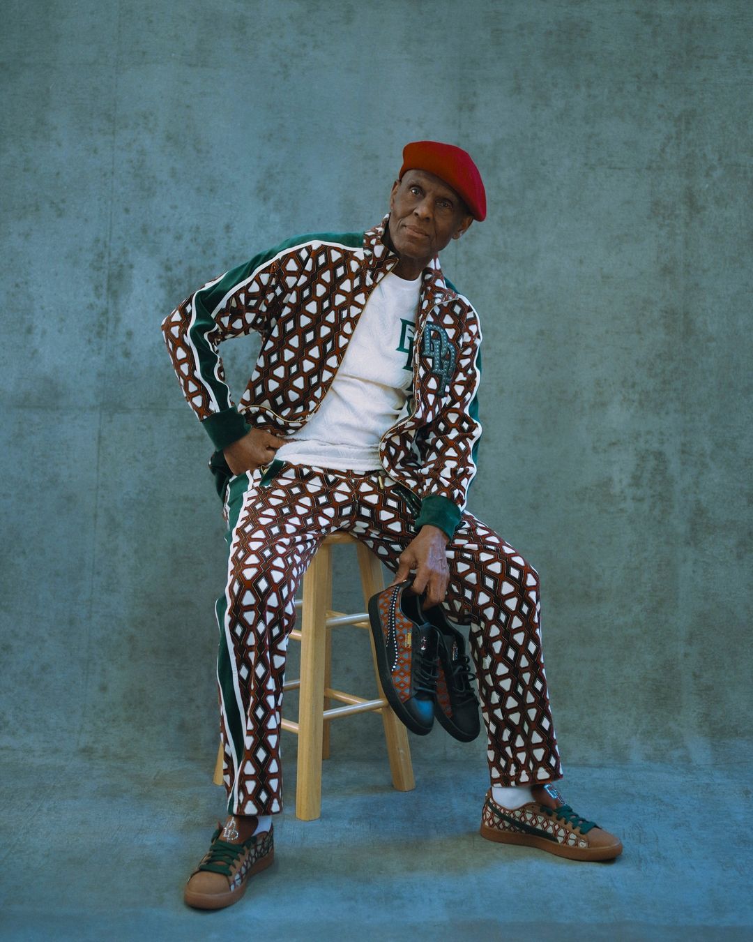Older Black man in patterned track suit and red beret sitting on a stool holding shoes in a photo studio with a grey background.