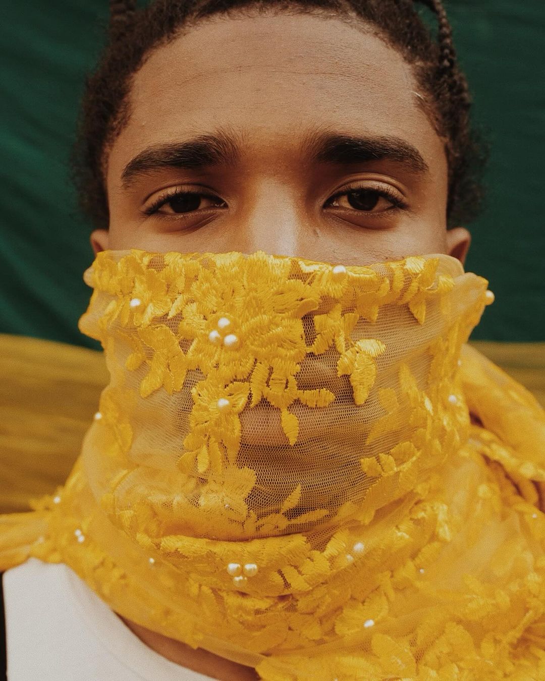 Close-up portrait of a young adult with braided hair partially concealed by a yellow floral lace veil in front of a green backdrop.