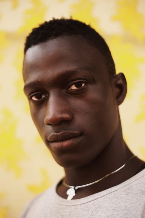 Close-up portrait of a young adult male with dark skin and short black hair, wearing a gray shirt and a silver necklace with a pendant.