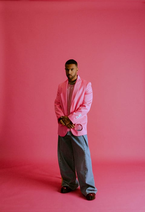 Man in a pink blazer and oversized jeans stands confidently against a vibrant pink backdrop in a full-length studio shot.