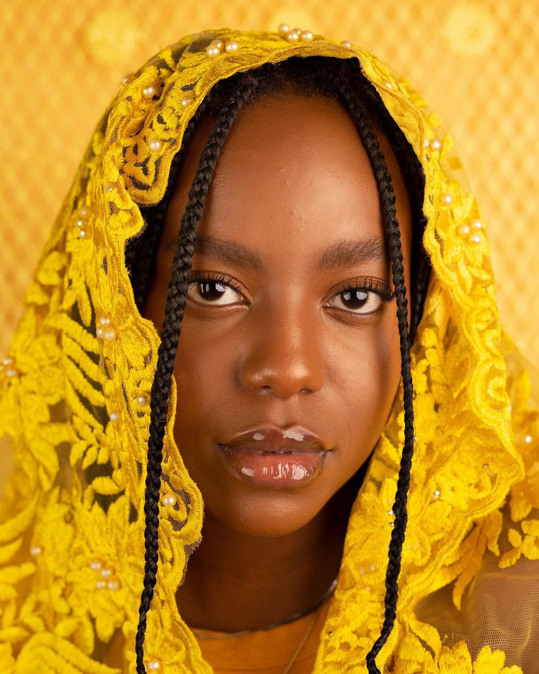 Close-up of an African-American female model with braided hair, wearing a yellow lace head covering with pearl details, against a yellow background.