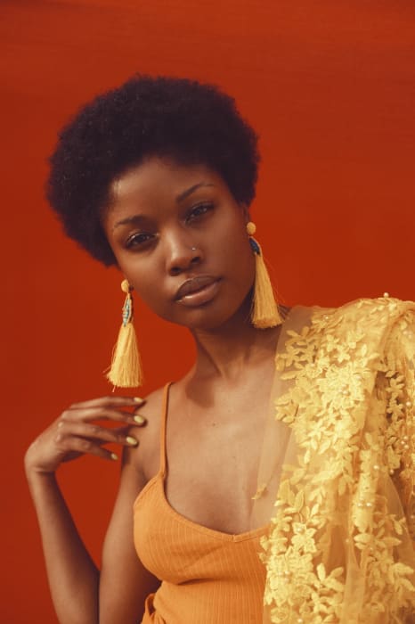Elegant portrait of an African American woman with an afro, wearing yellow earrings and a sheer floral shawl, against a red backdrop.