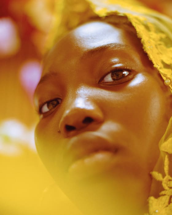 Close-up portrait of a young adult woman with brown eyes, wearing a yellow lace head covering, against a soft-focus background.