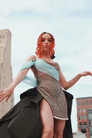 A stylish adult female with curly red hair, wearing a shimmering dress with a black train, poses in an urban setting with tall buildings.