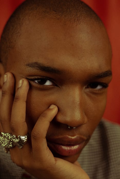 Close-up portrait of a Black man with a shaved head and septum ring resting his chin on a hand with a large silver ring.