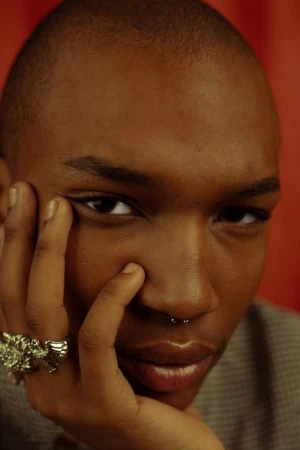 Close-up portrait of a Black man with a shaved head and septum ring resting his chin on a hand with a large silver ring.