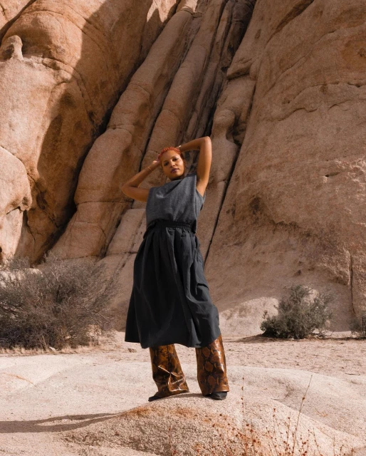 An adult woman in a sleeveless top and skirt over patterned pants stands in a desert landscape with rock formations and sparse vegetation.