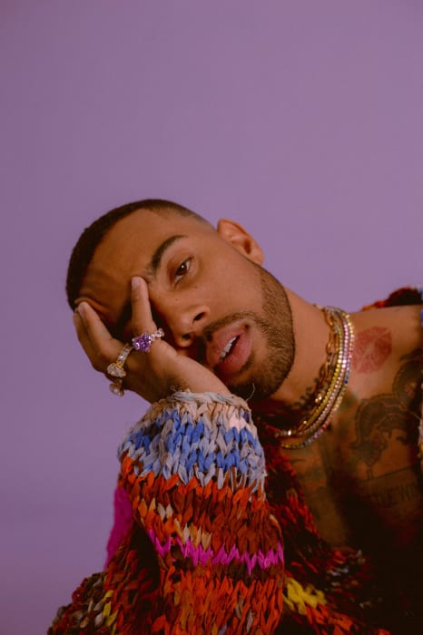 Close-up studio shot of a Black adult male with stubble, adorned with jewelry and a multicolored sweater against a light purple backdrop.