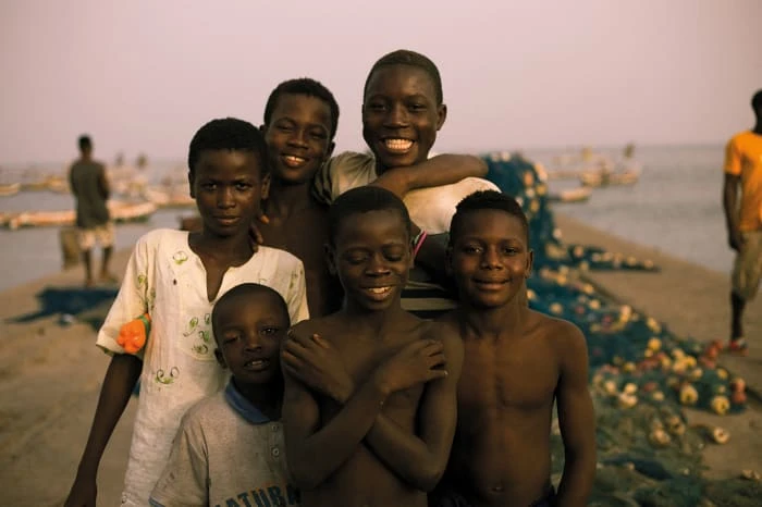 Group of African boys pose outdoors on a beach near fishing nets and boats, smiling and looking at the camera in soft lighting.