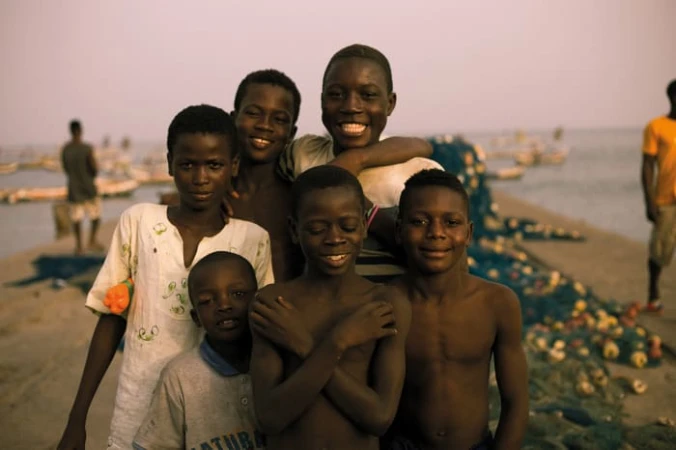 Group of African boys pose outdoors on a beach near fishing nets and boats, smiling and looking at the camera in soft lighting.
