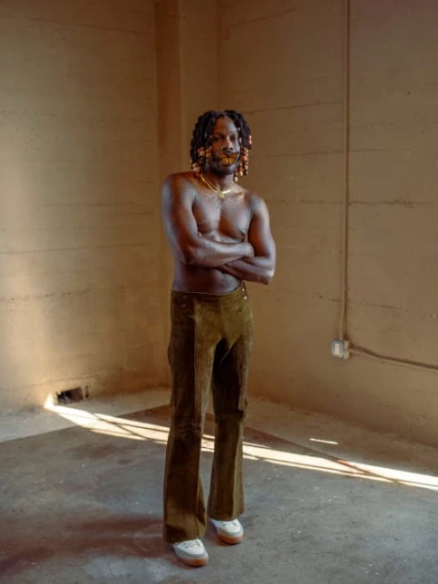 Shirtless Black man with dreadlocks and gold jewelry strikes a confident pose in olive green corduroy pants against a neutral indoor background.