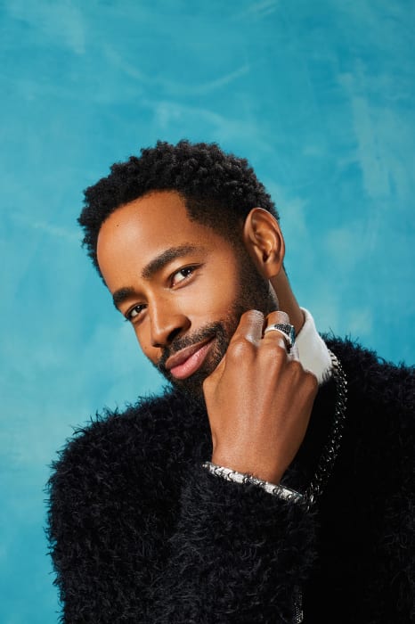 Portrait of a Black man with a beard wearing a black fuzzy sweater and a silver necklace against a mottled light blue background.