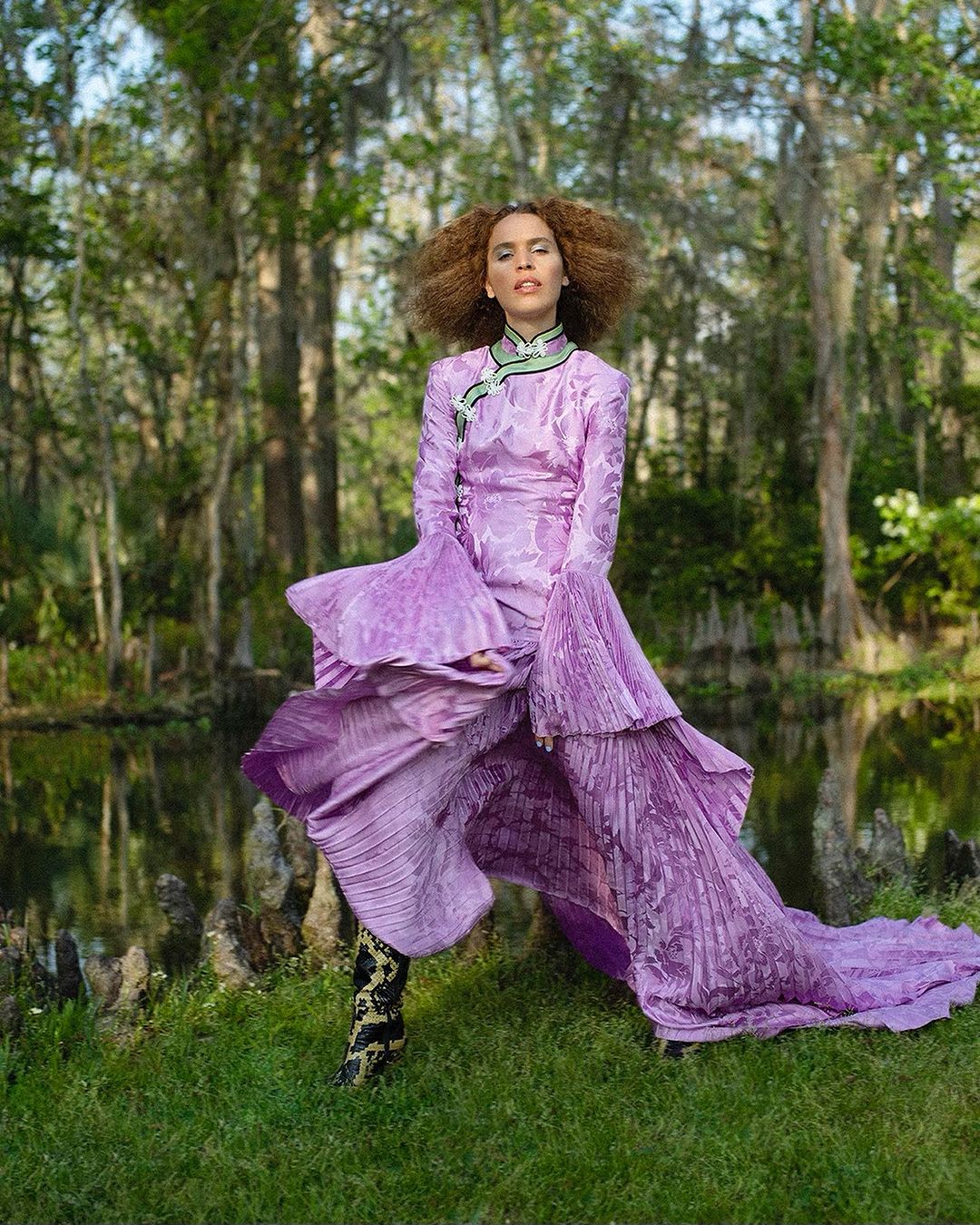 Adult female in a lavender gown with pleated details poses in a grassy marsh landscape with trees and a shallow body of water.