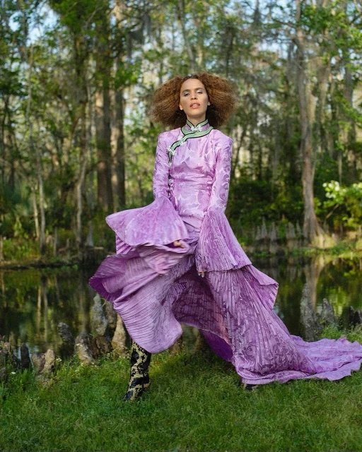 Adult female in a lavender gown with pleated details poses in a grassy marsh landscape with trees and a shallow body of water.