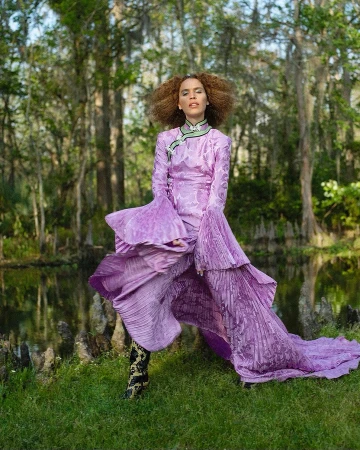 Adult female in a lavender gown with pleated details poses in a grassy marsh landscape with trees and a shallow body of water.