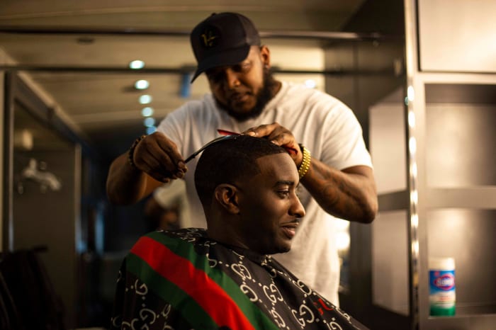 Man getting haircut from barber in indoor setting with Gucci print cape, wearing a black baseball cap, white t-shirt, and gold bracelet.