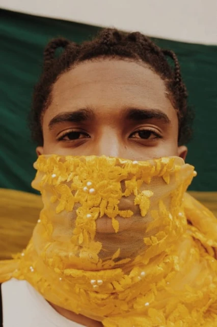 Close-up of a young adult with braided hair, partially obscured by an embroidered yellow lace scarf, set against a green background.