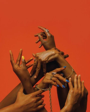 Multiple dark-skinned hands reach up, adorned with gold jewelry and colorful manicures, against a vibrant orange background.