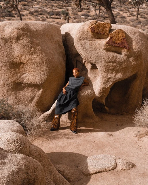 A woman in a sleeveless top and dark skirt poses among large rocks in a desert landscape with sparse trees, creating a stylish outdoor shot.