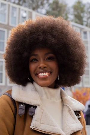 Portrait of a Black woman with a large afro hairstyle smiling while wearing a tan jacket with a white shearling collar