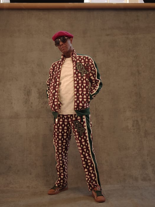Man in patterned tracksuit and beret poses indoors against a gray backdrop, creating a fashion-forward and stylish portrait.