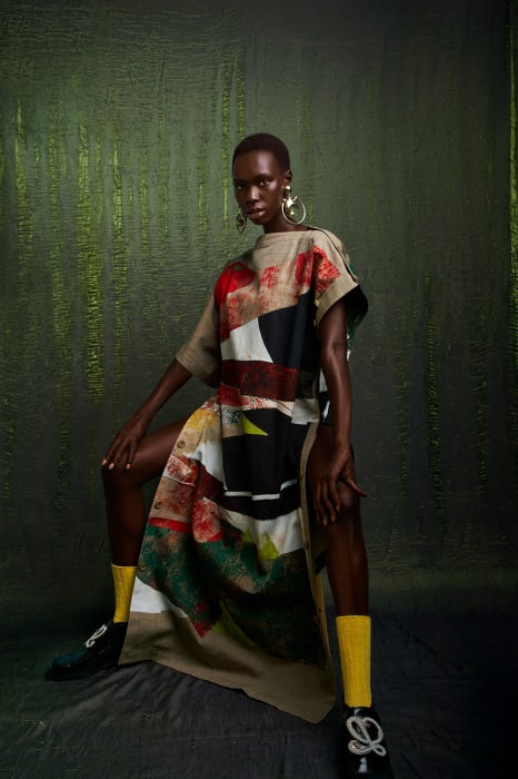 A dark-skinned woman poses in a flowing patterned dress with yellow socks and embellished shoes against a textured olive-green background.