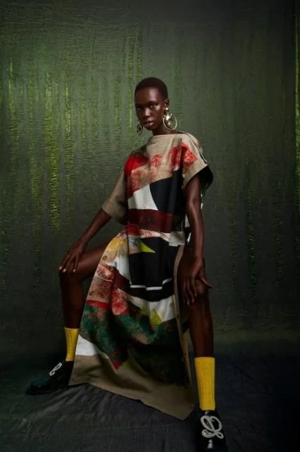 A dark-skinned woman poses in a flowing patterned dress with yellow socks and embellished shoes against a textured olive-green background.
