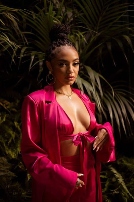 Portrait of a woman with dark hair and a topknot, wearing a pink satin suit, posing against a dark green tropical foliage backdrop.
