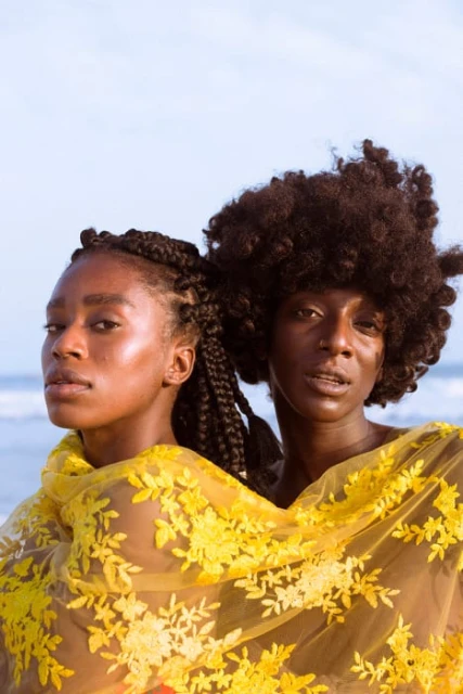 Two Black women with braided and afro hairstyles, wrapped in a yellow floral lace shawl, pose against a light blue sky and beach backdrop.