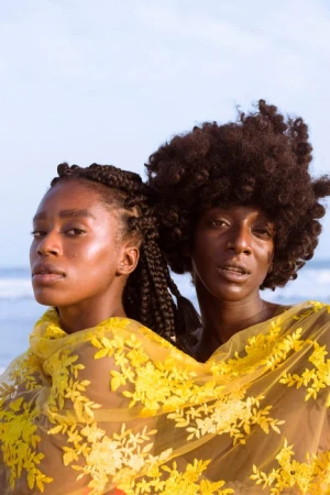 Two Black women with braided and afro hairstyles, wrapped in a yellow floral lace shawl, pose against a light blue sky and beach backdrop.