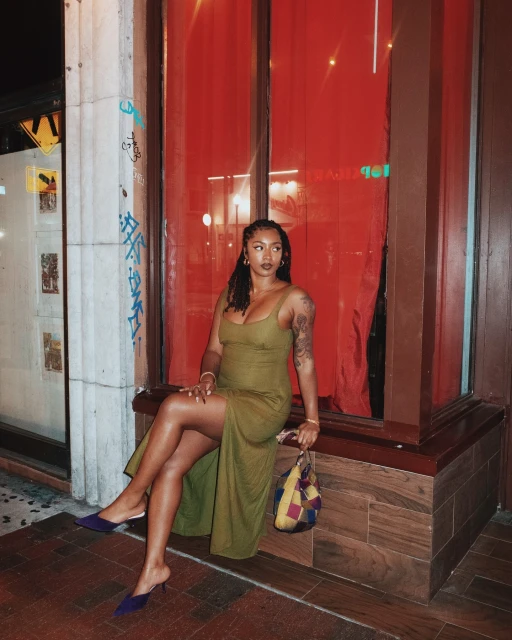 A woman in a dress sits on a ledge outside a building with a colorful purse and dress shoes, with a red backdrop and city environment.