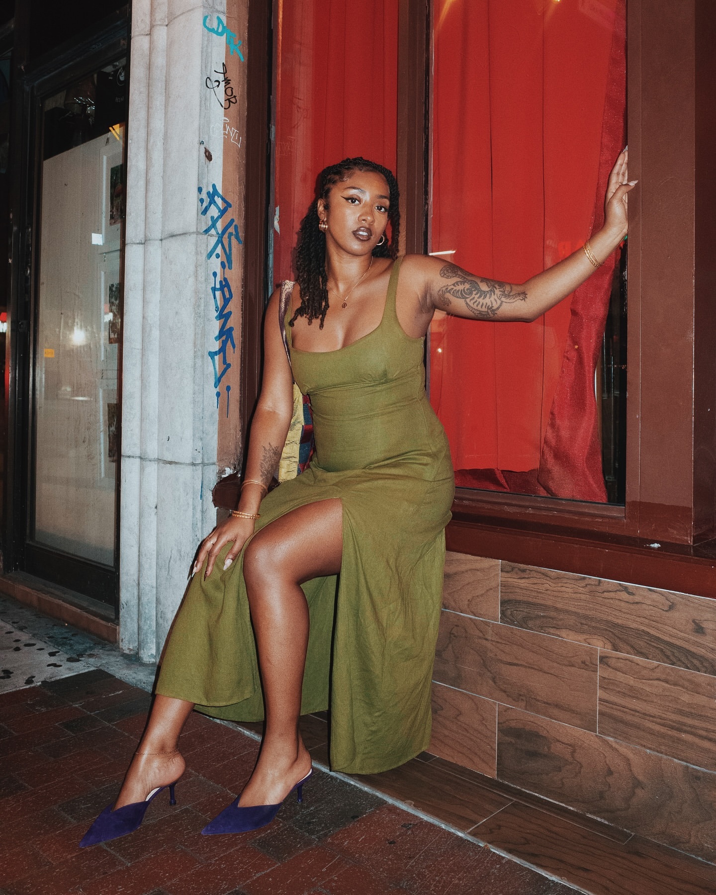 A young woman with tattoos, wearing an olive green dress and blue heels, poses in an urban setting with red accents in the evening.