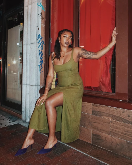 A young woman with tattoos, wearing an olive green dress and blue heels, poses in an urban setting with red accents in the evening.