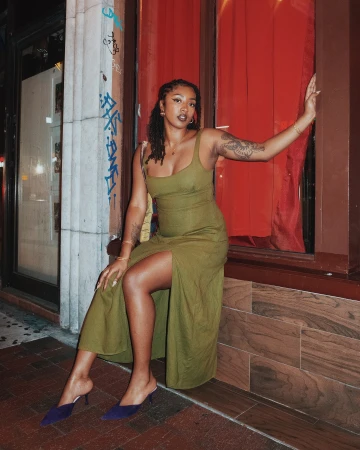 A young woman with tattoos, wearing an olive green dress and blue heels, poses in an urban setting with red accents in the evening.
