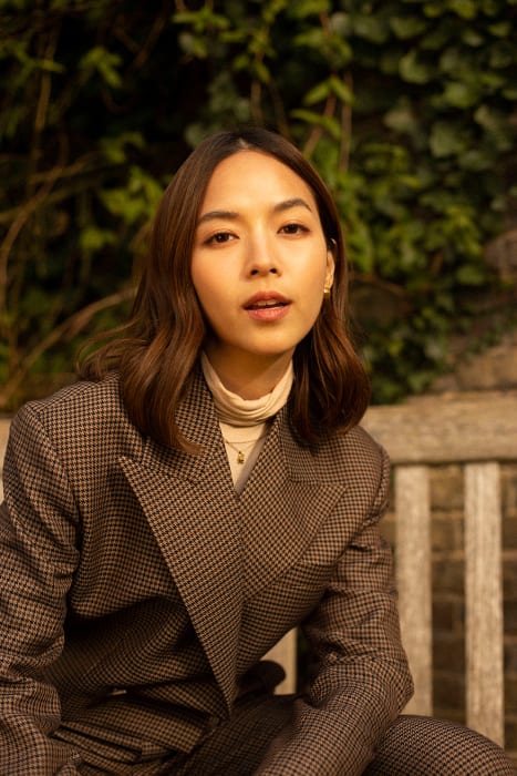 Portrait of a young woman with brown hair wearing a houndstooth blazer, sitting on a bench with greenery in the background.