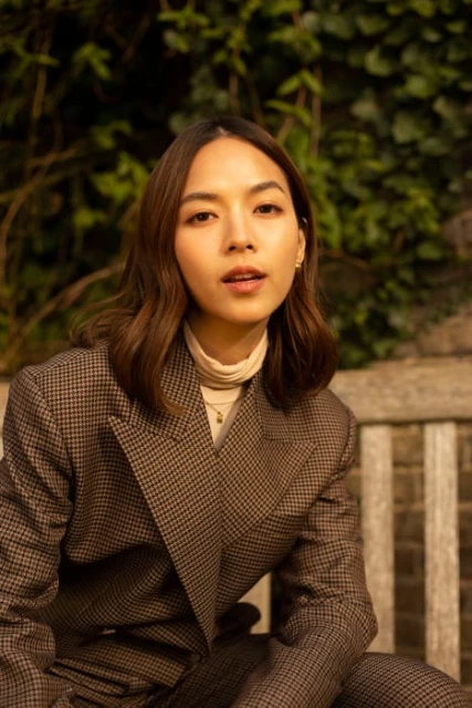 Portrait of a young woman with brown hair wearing a houndstooth blazer, sitting on a bench with greenery in the background.