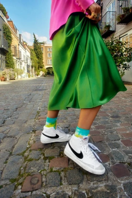 Person wearing pink top, green satin skirt, white Nike sneakers, and colorful socks on a cobblestone street lined with brick buildings.