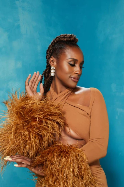 Portrait of adult female with light skin, brown hair, in long tan dress with feather boa sleeves, set against an aqua background.