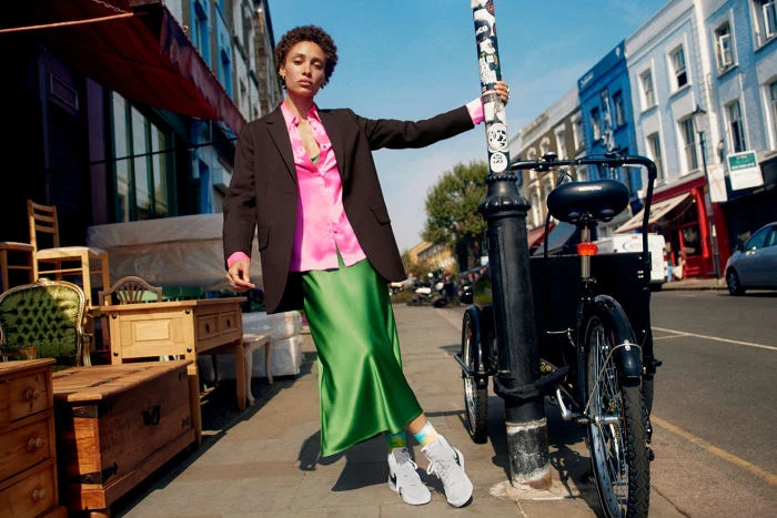 Woman in oversized blazer, pink shirt, and green skirt poses on a city sidewalk next to a bicycle; vibrant fashion and urban setting.
